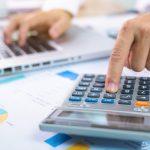 person with one hand typing on a laptop and the other working a calculator